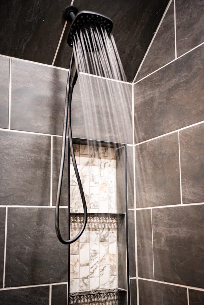 Matte black handheld showerhead spraying water over a tiled shower with decorative marble-look niches.